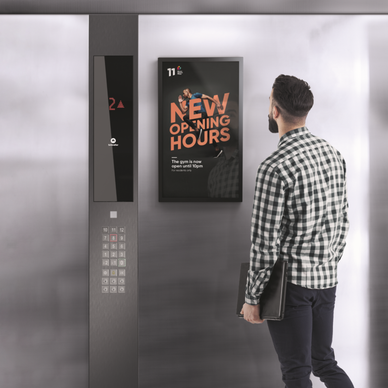 A man with short hair and a beard, wearing a black-and-white checkered shirt and dark pants, stands in an elevator and looks at a digital screen displaying an advertisement. The screen reads "NEW OPENING HOURS" in bold orange text, featuring an image of a man exercising. Below it, smaller text states, "The gym is now open until 10pm. For residents only." To the left of the screen is the elevator control panel with floor buttons and a small display showing the number "2" in red. The elevator interior has a sleek metallic finish.