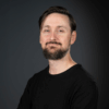 Portrait of Andrew Jarder, Chief Product Officer at Grassfish, standing confidently with arms crossed, wearing a black sweater against a dark background.