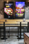 Interior of a modern burger restaurant featuring two digital menu screens. The left screen displays a close-up of a double cheeseburger with the text 'EXTRA HUNGRIG? BESTÄLL DUBBELT KÖTT +40 KR' from Bastard Burgers. The right screen showcases a vibrant burger with purple toppings, labeled 'JÄVLIGT KOREA,' with a lightning graphic. The setting includes wooden tables, black metal stools, and a tiled wall with graffiti-style artwork.