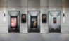 A modern elevator lobby featuring three elevator doors set within a sleek gray stone wall. Each elevator displays digital signage: the left elevator shows a pink advertisement for perfume with text reading 
