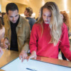 A young woman in a red sweater interacts with a large touchscreen table while a man in a tan jacket watches and points at the screen. They are in a retail or showroom environment with other people socializing in the background, some holding drinks.