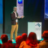 A speaker stands on stage at the GRASSFISH Summit 2025, gesturing with his hands while addressing an audience. Behind him is a large screen displaying an image of an old Samsung mobile phone. The speaker wears a dark jacket, white shirt, mustard-colored pants, and white sneakers. A podium with the summit logo is placed nearby, and attendees are seated facing the stage.