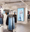 Modern clothing store interior with bright lighting. Prominent digital in-store retail media screen displays an advertisement featuring a smiling woman in floral attire. Denim jeans are neatly hung on racks nearby, creating a casual, inviting shopping ambiance.