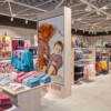 The interior of a Lindex store, specifically the children’s clothing section. The store features a modern design with a light-colored tiled floor and a grid ceiling with spotlights. In the foreground, a wooden display table presents neatly folded kids’ clothes in pink, blue, and red tones, along with a sign indicating a 20% discount. To the left, racks display vibrant children’s outfits, including dresses and jackets. A central freestanding digital screen showcases an image of a toddler sitting next to a large teddy bear, adding a cozy and playful element to the store's ambiance. The background includes additional racks with organized clothing items such as sweaters and pants, emphasizing the store's focus on stylish and practical kidswear.