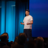 A person wearing a white T-shirt, dark pants, and a cap stands on a stage giving a presentation. They hold a small device in one hand while addressing an audience seated in front of them. The stage is lit with blue lighting and features a large screen in the background.