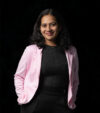 A smiling woman in a pink blazer and black outfit stands confidently against a dark background, conveying a professional and approachable tone.