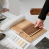 A hand selecting a wooden material sample from a collection of interior design materials. The workspace includes wood swatches, a fur textile sample, a color palette with neutral and warm tones, and a dark marble tile. The setting suggests a design or renovation planning process.