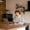 A young woman with short dark hair, wearing a cozy gray sweater, sits at a wooden kitchen counter, writing in a notebook while using her laptop. The modern kitchen features white subway tiles, wooden shelves with potted plants, a black range hood, and a stylish black refrigerator in the background, creating a warm and contemporary home office atmosphere.