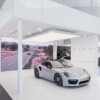 A silver Porsche sports car displayed in a modern showroom with a sleek white interior. A large digital screen in the background shows a scenic road with cars driving through a countryside landscape.