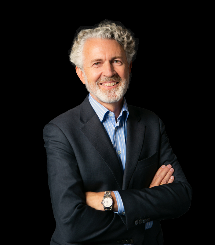 Smiling man with curly gray hair and a beard wearing a dark suit and blue striped shirt, arms crossed. The background is plain black, conveying confidence.