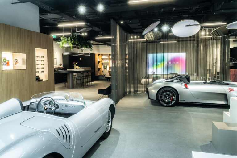 A luxurious Porsche showroom featuring two classic silver Porsche sports cars. The space includes modern lighting, a lounge area with a bar, bookshelves, and a display of Porsche-branded sunglasses. A digital screen in the background displays the Porsche logo with a colorful gradient.
