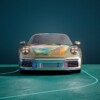 A Porsche sports car with a gold exterior, featuring a colorful digital projection on the hood. The car is positioned on a teal floor with curved light accents, against a dark teal background.