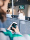 A person holding a smartphone and interacting with a digital interface featuring hexagonal elements on the screen. The setting appears to be a public or educational space with two young girls in the background near a railing, and a digital display screen mounted on the wall.
