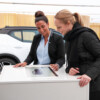 Two women engaged in an interactive car configuration experience at a digital touchscreen kiosk in a showroom. One woman, wearing a Volvo-branded uniform, is demonstrating the customization options on the screen, while the other, dressed in a black winter coat, smiles as they explore vehicle features together. A white Volvo SUV is visible in the background.