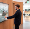 A man in a dark suit interacts with a large touchscreen display in a modern showroom with wooden paneling. The screen reflects a Volvo showroom environment, with a white Volvo XC60 visible in the background. The setting is well-lit, with natural light coming through large windows, enhancing the sleek and minimalistic interior design.