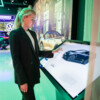 A woman interacts with a sleek digital touchscreen display showcasing Volkswagen vehicles in a modern showroom. The screen highlights detailed information about a black Volkswagen car, including its price and features. In the background, illuminated branding and visuals of other Volkswagen models enhance the immersive experience.