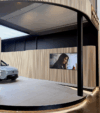 A sleek, modern car is displayed partially on a rotating platform beside a textured wooden wall. A screen shows a smiling person, evoking a warm, inviting tone.